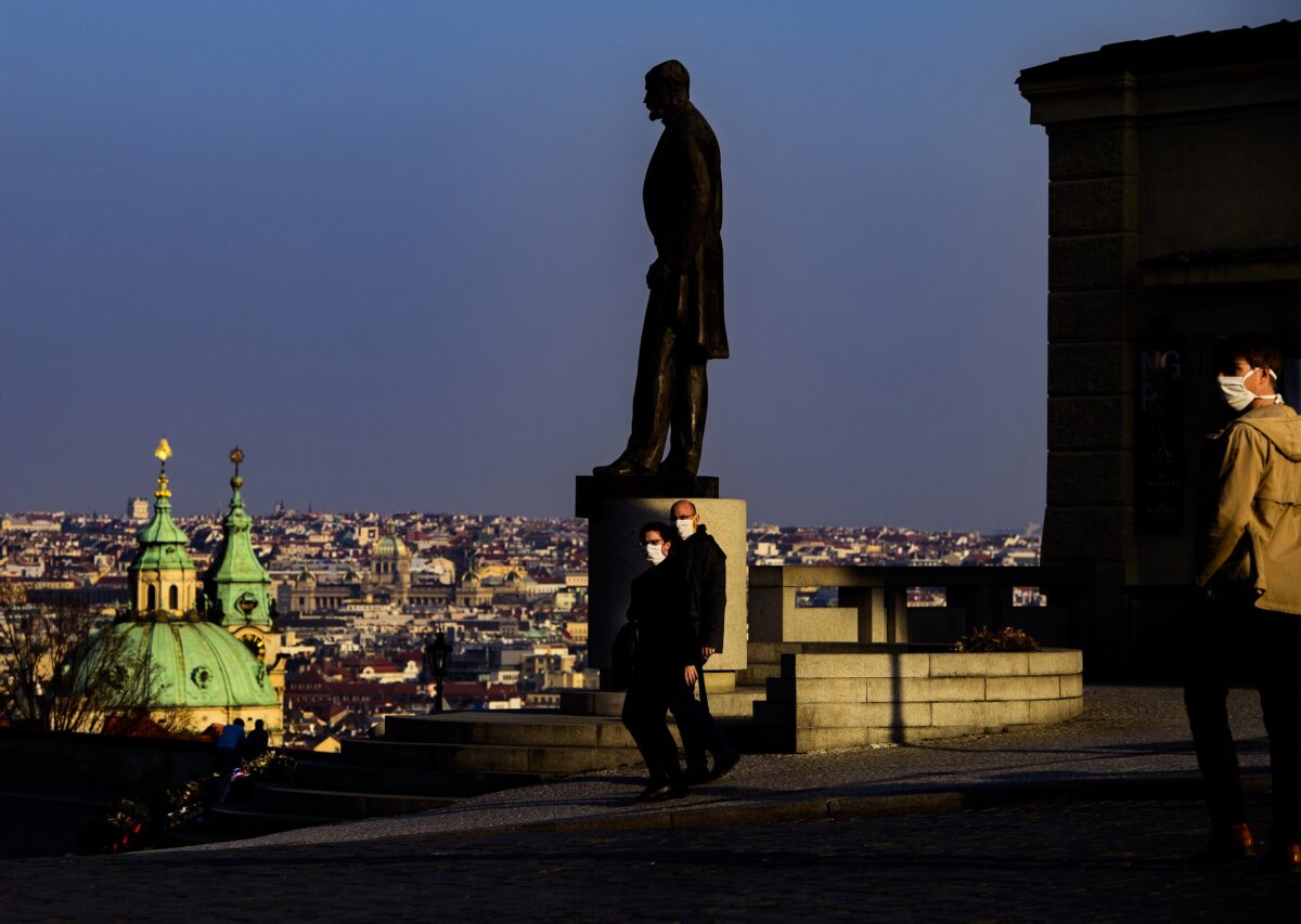 Pohled na sochu Tomáše Masaryka na pozadí večerní Prahy, kolem níž procházejí lidé v rouškách